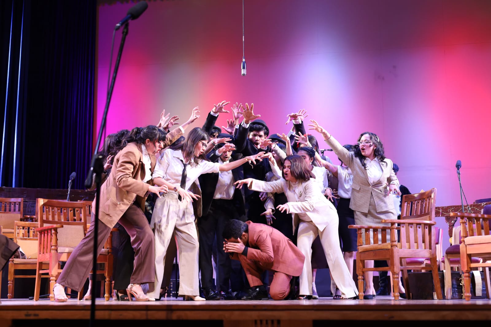 Trial by Jury ensemble performing a lively group scene on stage, with actors in formal costumes under vibrant pink and purple lighting.