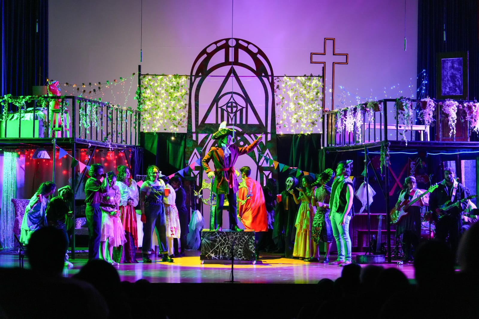 Romeo & Juliet ensemble performing on stage under vibrant pink and purple lighting, with actors gathered in a dramatic group scene.